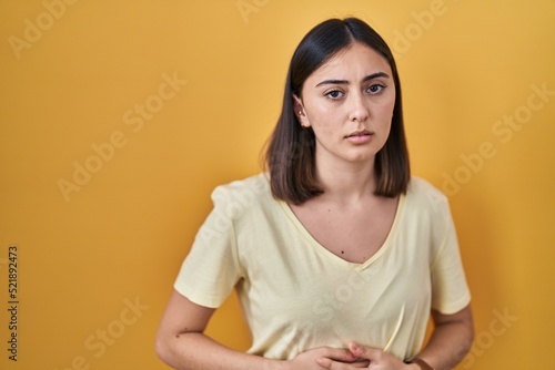 Hispanic girl wearing casual t shirt over yellow background with hand on stomach because indigestion, painful illness feeling unwell. ache concept.
