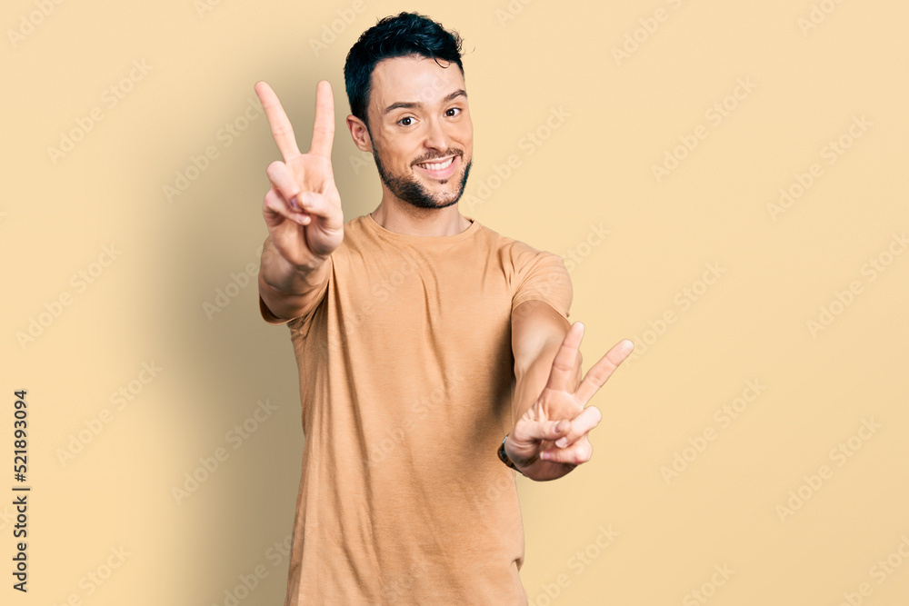 Hispanic man with beard wearing casual t shirt smiling with tongue out showing fingers of both hands doing victory sign. number two.