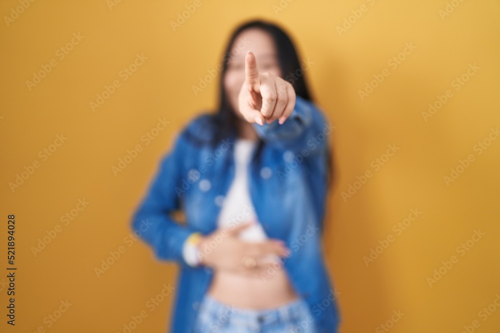 Young asian woman standing over yellow background laughing at you, pointing finger to the camera with hand over body, shame expression