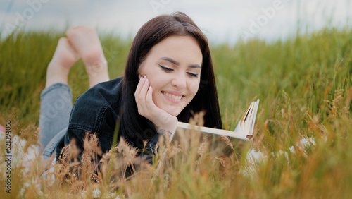 Young serene caucasian brunet woman girl lying on grass outdoors attractive smiling teenage student reads book enjoy romantic literature history relax outside spend time cultural leisure lazy weekends