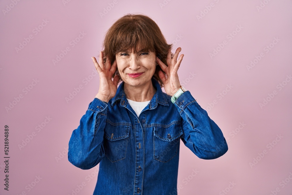 Middle age woman standing over pink background covering ears with fingers with annoyed expression for the noise of loud music. deaf concept. Middle age woman standing over pink background covering ears with fingers with annoyed expression for the noise of loud music. deaf concept.