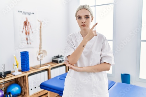 Young caucasian woman working at pain recovery clinic pointing with hand finger to the side showing advertisement  serious and calm face