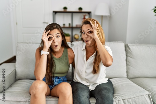 Mother and daughter together sitting on the sofa at home doing ok gesture shocked with surprised face, eye looking through fingers. unbelieving expression.