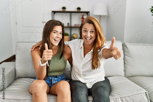 Mother and daughter together sitting on the sofa at home approving doing positive gesture with hand, thumbs up smiling and happy for success. winner gesture.