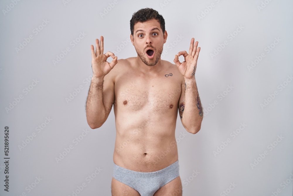 Fototapeta premium Young hispanic man standing shirtless wearing underware looking surprised and shocked doing ok approval symbol with fingers. crazy expression