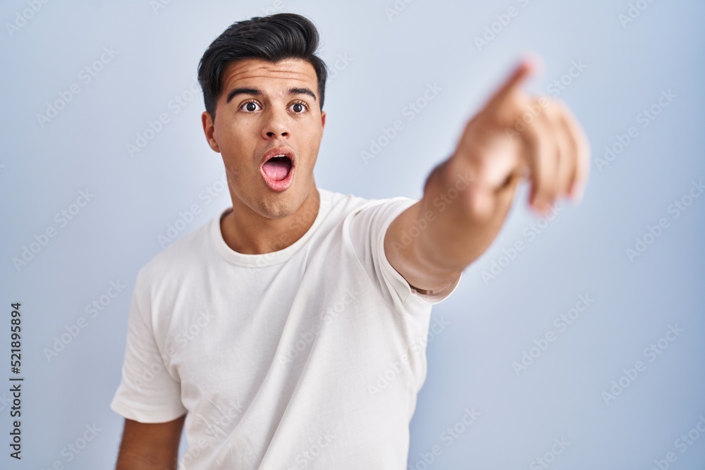 Hispanic man standing over blue background pointing with finger surprised ahead, open mouth amazed expression, something on the front Hispanic man standing over blue background pointing with finger surprised ahead, open mouth amazed expression, something on the front