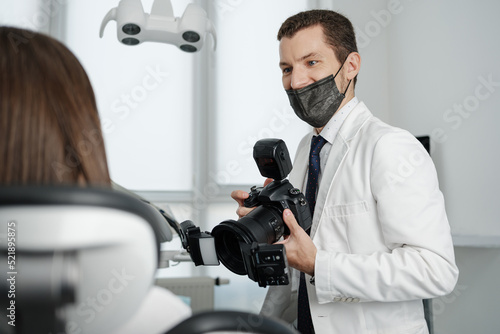 Handsome dentist making photo of female patient's smile after treatment. Special camera with flash ring shadowless.