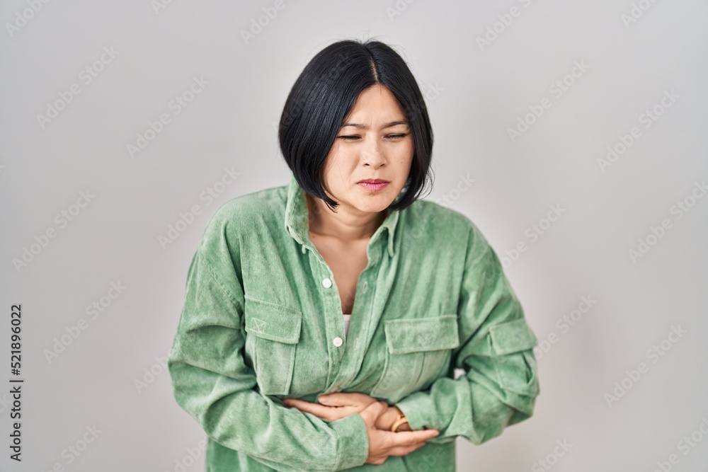 Young asian woman standing over white background with hand on stomach because indigestion, painful illness feeling unwell. ache concept. Young asian woman standing over white background with hand on stomach because indigestion, painful illness feeling unwell. ache concept.