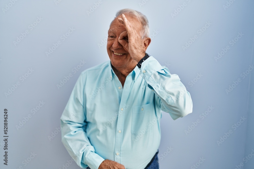 Senior man with grey hair standing over blue background doing ok gesture with hand smiling, eye looking through fingers with happy face. Senior man with grey hair standing over blue background doing ok gesture with hand smiling, eye looking through fingers with happy face.