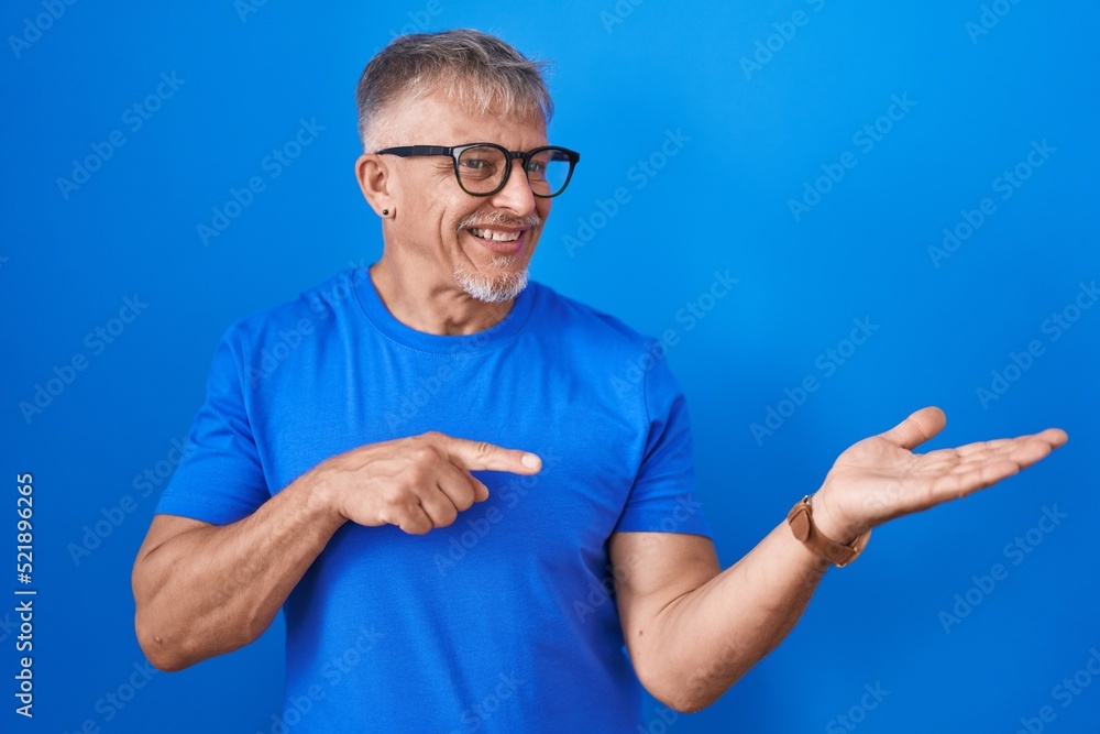 Hispanic man with grey hair standing over blue background amazed and smiling to the camera while presenting with hand and pointing with finger. Hispanic man with grey hair standing over blue background amazed and smiling to the camera while presenting with hand and pointing with finger.