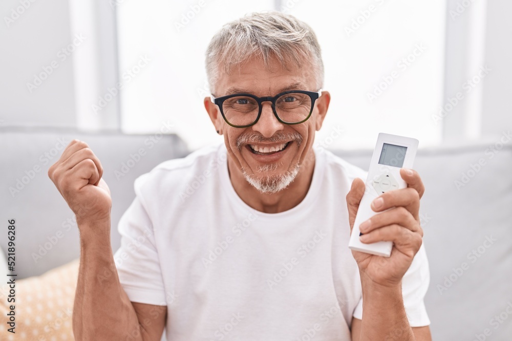 Hispanic man with grey hair holding air conditioner control screaming proud, celebrating victory and success very excited with raised arm