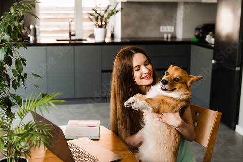 Girl with laptop smile and play with Corgi dog at home. Programmer woman working online and have a break for hug her dog. Having fun with Welsh Corgi Pembroke.