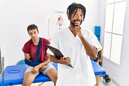 Young hispanic man working at pain recovery clinic with a man with broken arm with hand on chin thinking about question  pensive expression. smiling and thoughtful face. doubt concept.