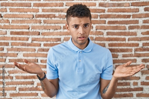 Brazilian young man standing over brick wall clueless and confused with open arms, no idea concept.
