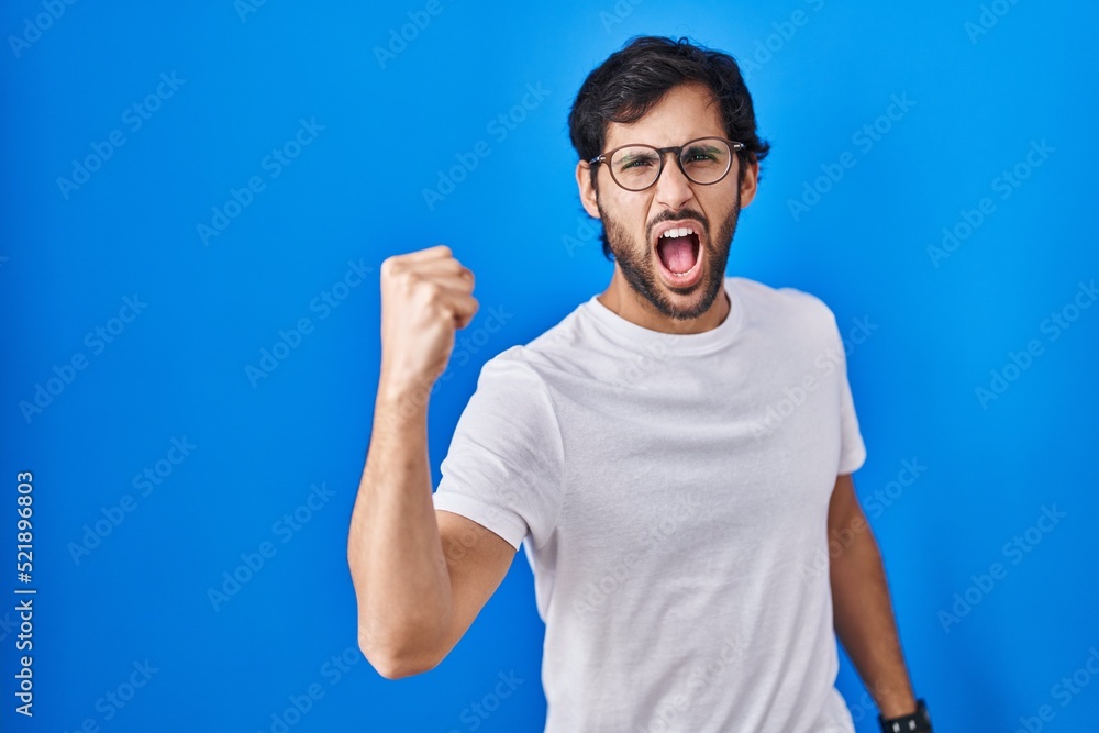 Handsome latin man standing over blue background angry and mad raising fist frustrated and furious while shouting with anger. rage and aggressive concept. Handsome latin man standing over blue background angry and mad raising fist frustrated and furious while shouting with anger. rage and aggressive concept.