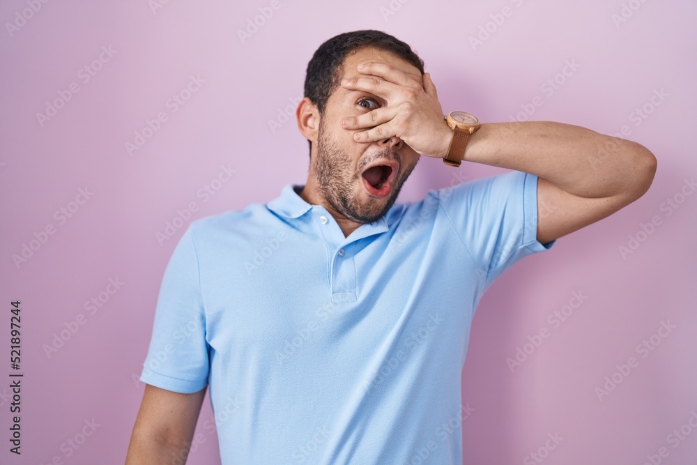 Hispanic man standing over pink background peeking in shock covering face and eyes with hand, looking through fingers with embarrassed expression. Hispanic man standing over pink background peeking in shock covering face and eyes with hand, looking through fingers with embarrassed expression.
