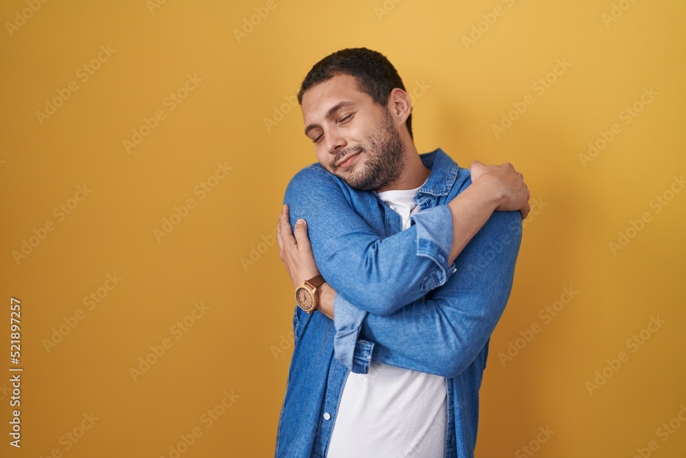 Hispanic man standing over yellow background hugging oneself happy and positive, smiling confident. self love and self care Hispanic man standing over yellow background hugging oneself happy and positive, smiling confident. self love and self care