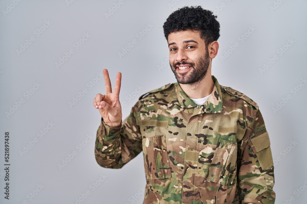 Arab man wearing camouflage army uniform smiling looking to the camera showing fingers doing victory sign. number two. Arab man wearing camouflage army uniform smiling looking to the camera showing fingers doing victory sign. number two.