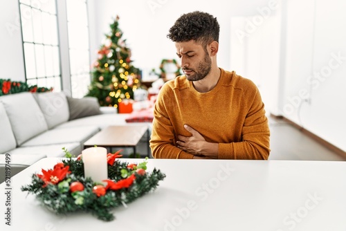 Arab young man sitting on the table by christmas tree with hand on stomach because nausea  painful disease feeling unwell. ache concept.