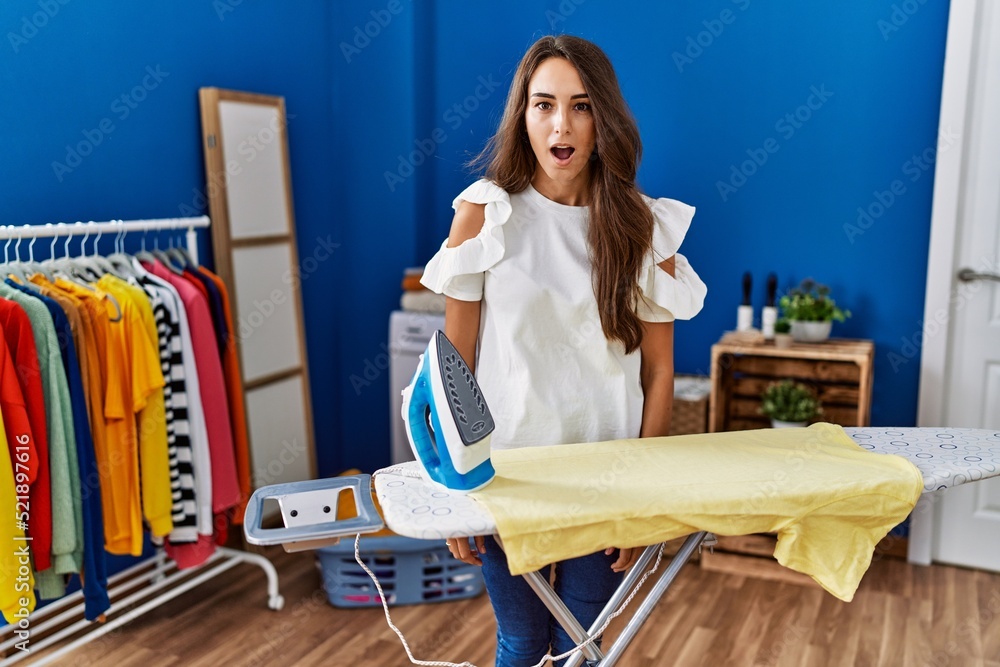 Young hispanic woman ironing clothes at laundry room afraid and shocked with surprise expression, fear and excited face. Young hispanic woman ironing clothes at laundry room afraid and shocked with surprise expression, fear and excited face.