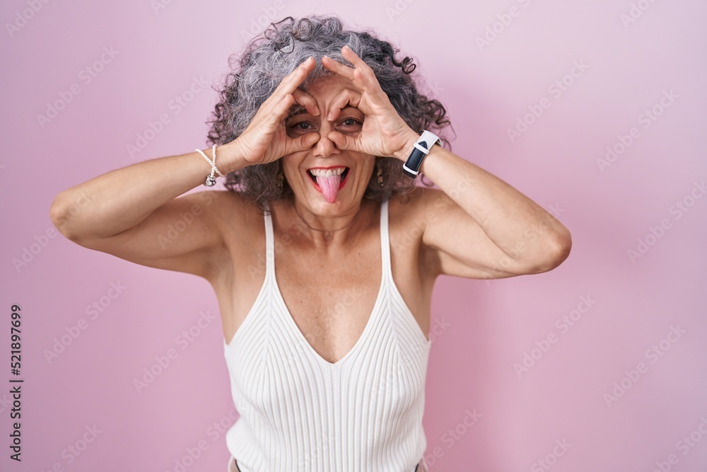 Middle age woman with grey hair standing over pink background doing ok gesture like binoculars sticking tongue out, eyes looking through fingers. crazy expression.