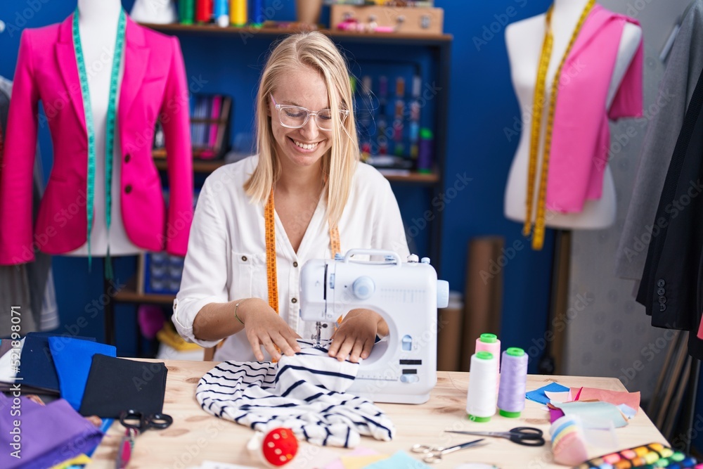 Fototapeta premium Young blonde woman tailor smiling confident using sewing machine at sewing studio
