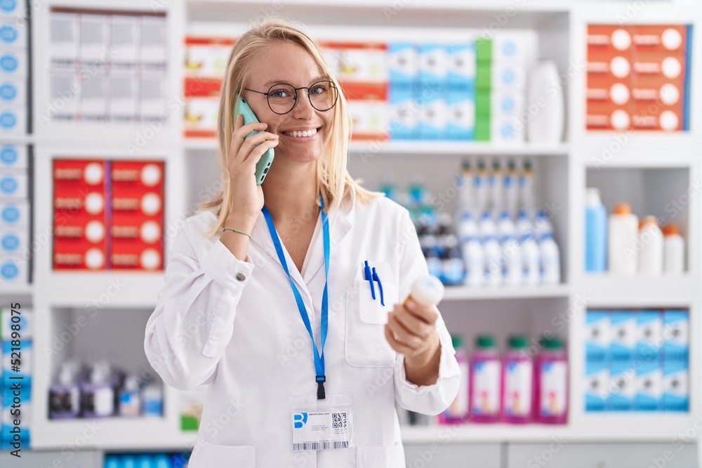 Young blonde woman pharmacist holding pills bottle talking on smartphone at pharmacy