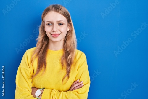 Young caucasian woman standing over blue background happy face smiling with crossed arms looking at the camera. positive person.