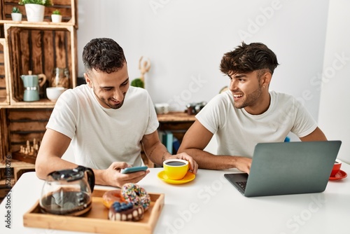 Two hispanic men couple having breakfast using smartphone and laptop at home