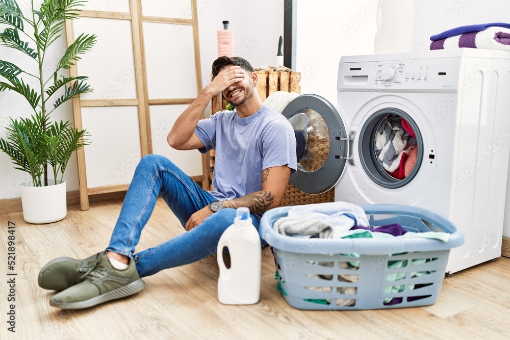 Young hispanic man putting dirty laundry into washing machine smiling and laughing with hand on face covering eyes for surprise. blind concept. Young hispanic man putting dirty laundry into washing machine smiling and laughing with hand on face covering eyes for surprise. blind concept.