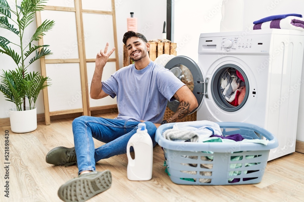 Young hispanic man putting dirty laundry into washing machine smiling looking to the camera showing fingers doing victory sign. number two. Young hispanic man putting dirty laundry into washing machine smiling looking to the camera showing fingers doing victory sign. number two.