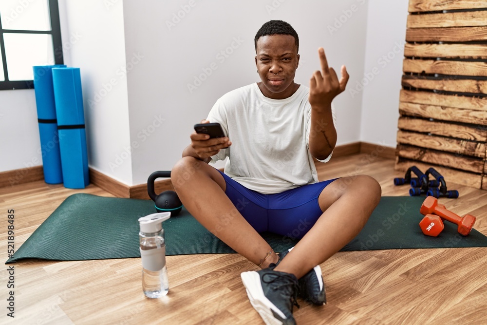 Young african man sitting on training mat at the gym using smartphone showing middle finger, impolite and rude fuck off expression Young african man sitting on training mat at the gym using smartphone showing middle finger, impolite and rude fuck off expression