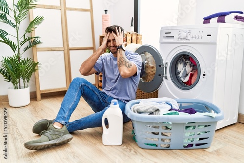 Obraz na plátně Young hispanic man putting dirty laundry into washing machine suffering from headache desperate and stressed because pain and migraine