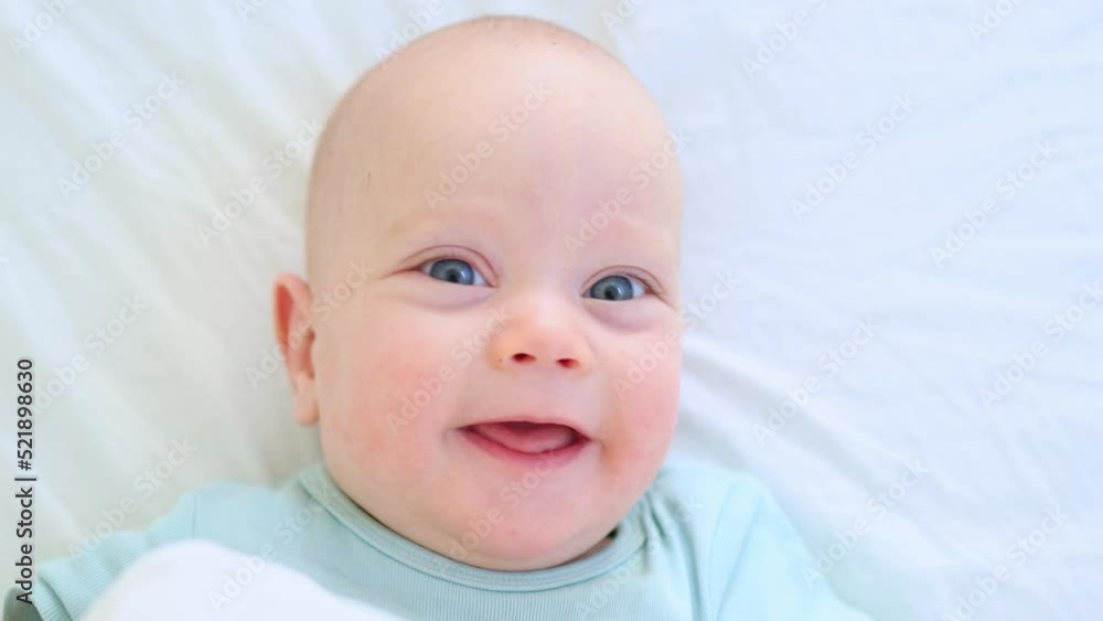 beautiful blue eyed infant baby portrait close-up, smiling face of little child lying in white bed on stomach. happy caucasian toddler baby portrait, funny kid looking camera opening mouth, emotions beautiful blue eyed infant baby portrait close-up, smiling face of little child lying in white bed on stomach. happy caucasian toddler baby portrait, funny kid looking camera opening mouth, emotions
