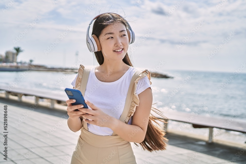 Young chinese girl smiling happy using headphones and smartphone at the promenade.