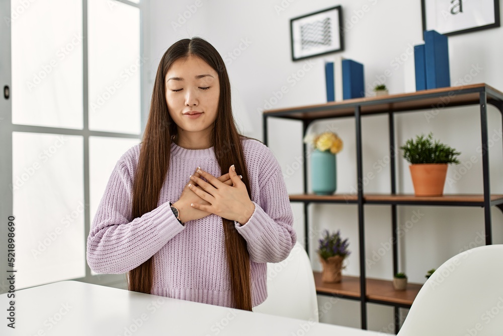 Young chinese girl wearing casual clothes sitting on the table at home smiling with hands on chest with closed eyes and grateful gesture on face. health concept. Young chinese girl wearing casual clothes sitting on the table at home smiling with hands on chest with closed eyes and grateful gesture on face. health concept.