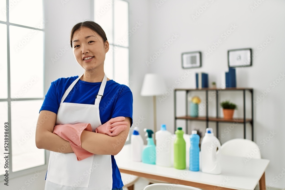 Young chinese housewife standing with arms crossed gesture and cleaning products on the table at home. Young chinese housewife standing with arms crossed gesture and cleaning products on the table at home.