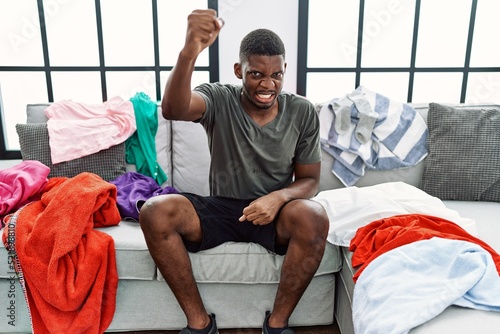 Young african american man sitting on the sofa with dirty laundry clothes angry and mad raising fist frustrated and furious while shouting with anger. rage and aggressive concept.