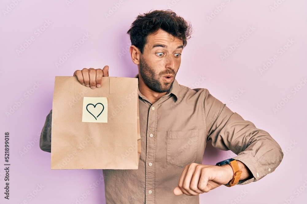 Handsome man with beard holding delivery paper bag with heart reminder looking at the watch time worried, afraid of getting late Handsome man with beard holding delivery paper bag with heart reminder looking at the watch time worried, afraid of getting late