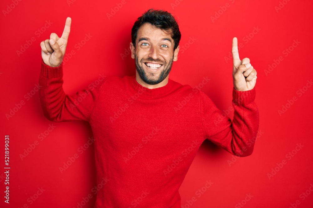 Handsome man with beard wearing casual red sweater smiling amazed and surprised and pointing up with fingers and raised arms. Handsome man with beard wearing casual red sweater smiling amazed and surprised and pointing up with fingers and raised arms.