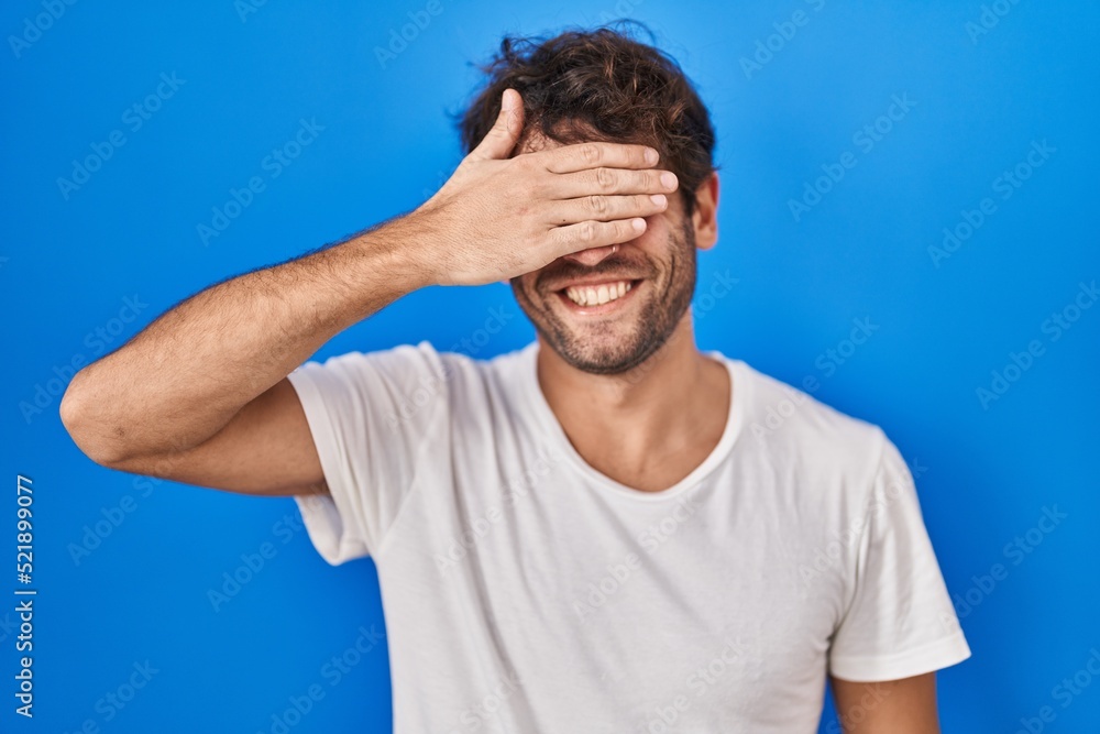 Hispanic young man standing over blue background smiling and laughing with hand on face covering eyes for surprise. blind concept. Hispanic young man standing over blue background smiling and laughing with hand on face covering eyes for surprise. blind concept.