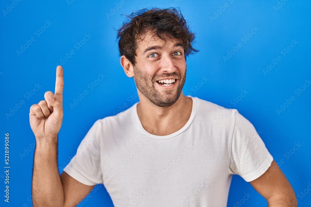 Hispanic young man standing over blue background pointing finger up with successful idea. exited and happy. number one. Hispanic young man standing over blue background pointing finger up with successful idea. exited and happy. number one.