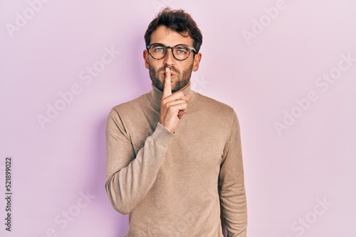 Handsome man with beard wearing turtleneck sweater and glasses asking to be quiet with finger on lips. silence and secret concept.
