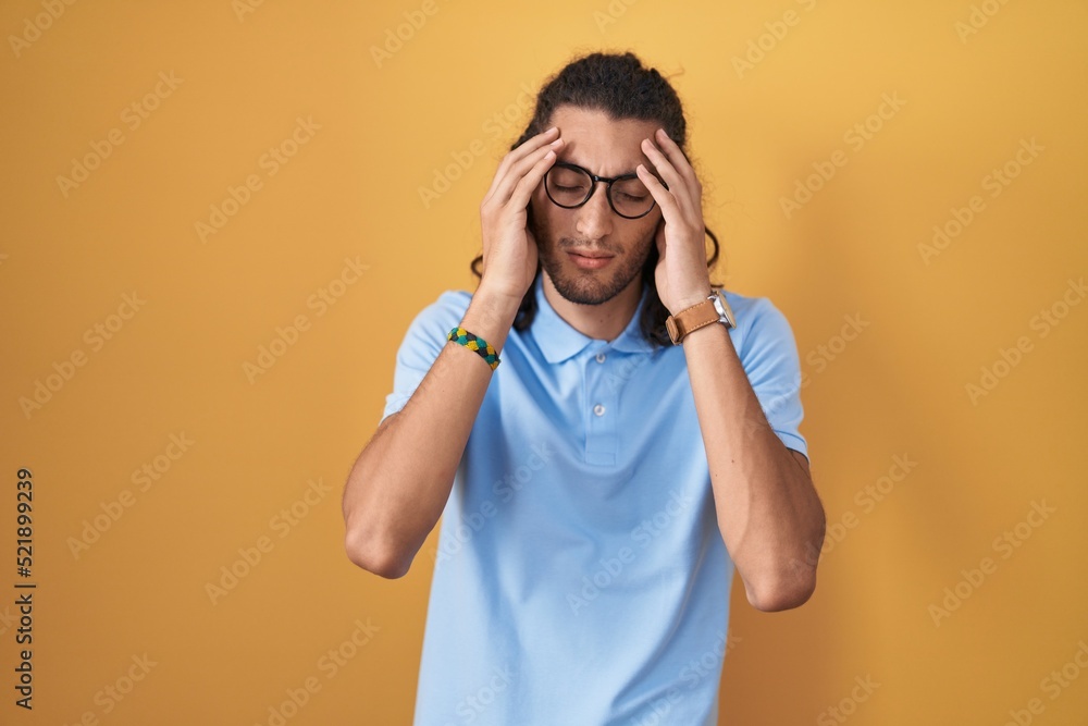 Young hispanic man standing over yellow background with hand on head, headache because stress. suffering migraine. Young hispanic man standing over yellow background with hand on head, headache because stress. suffering migraine.
