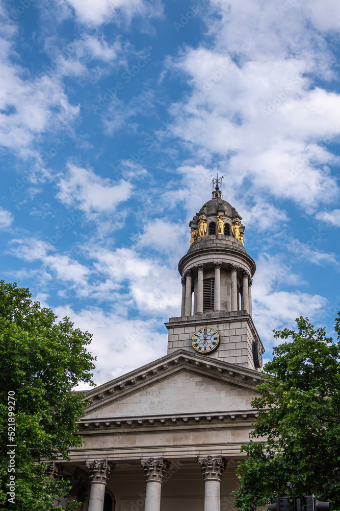 London, Great Britain - July 3, 2022: Closeup of clock tower with golden caryatid statues and pediment on columns of St. Marylebone Parish Church under blue cloudscape. Green foliage 