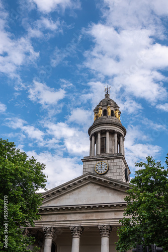 London, Great Britain - July 3, 2022: Closeup of clock tower with golden caryatid statues and pediment on columns of St. Marylebone Parish Church under blue cloudscape. Green foliage 