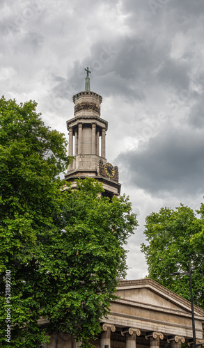 London, Great Britain - July 3, 2022: Beige stone tower and pediment above columns of St. Pancras New Church against heavy dark gray cloudscape and partly hidden by green foliage. photo