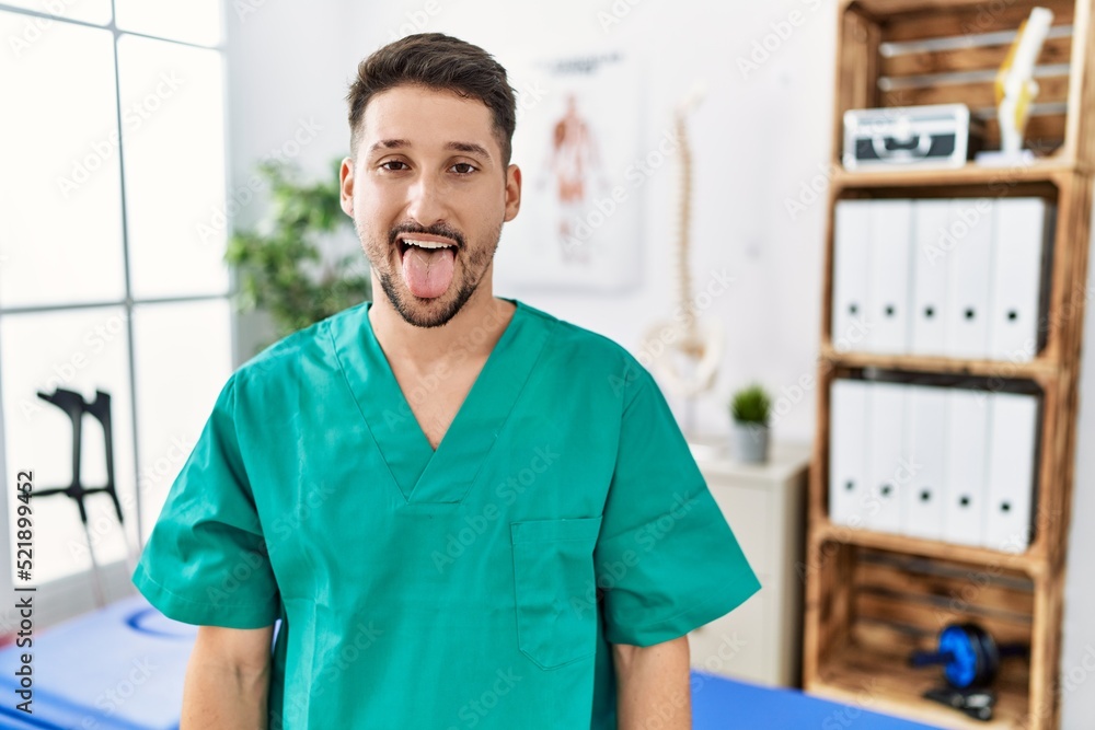 Young physiotherapist man working at pain recovery clinic sticking tongue out happy with funny expression. emotion concept. Young physiotherapist man working at pain recovery clinic sticking tongue out happy with funny expression. emotion concept.