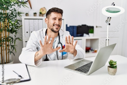 Young doctor working at the clinic using computer laptop afraid and terrified with fear expression stop gesture with hands, shouting in shock. panic concept.