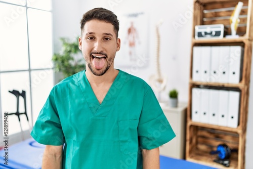 Young physiotherapist man working at pain recovery clinic sticking tongue out happy with funny expression. emotion concept.